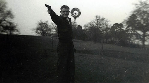 Staff Sgt Nealms stands on a beautiful hillside pointing his pistol. A windmill can be seen at the top of the hill.